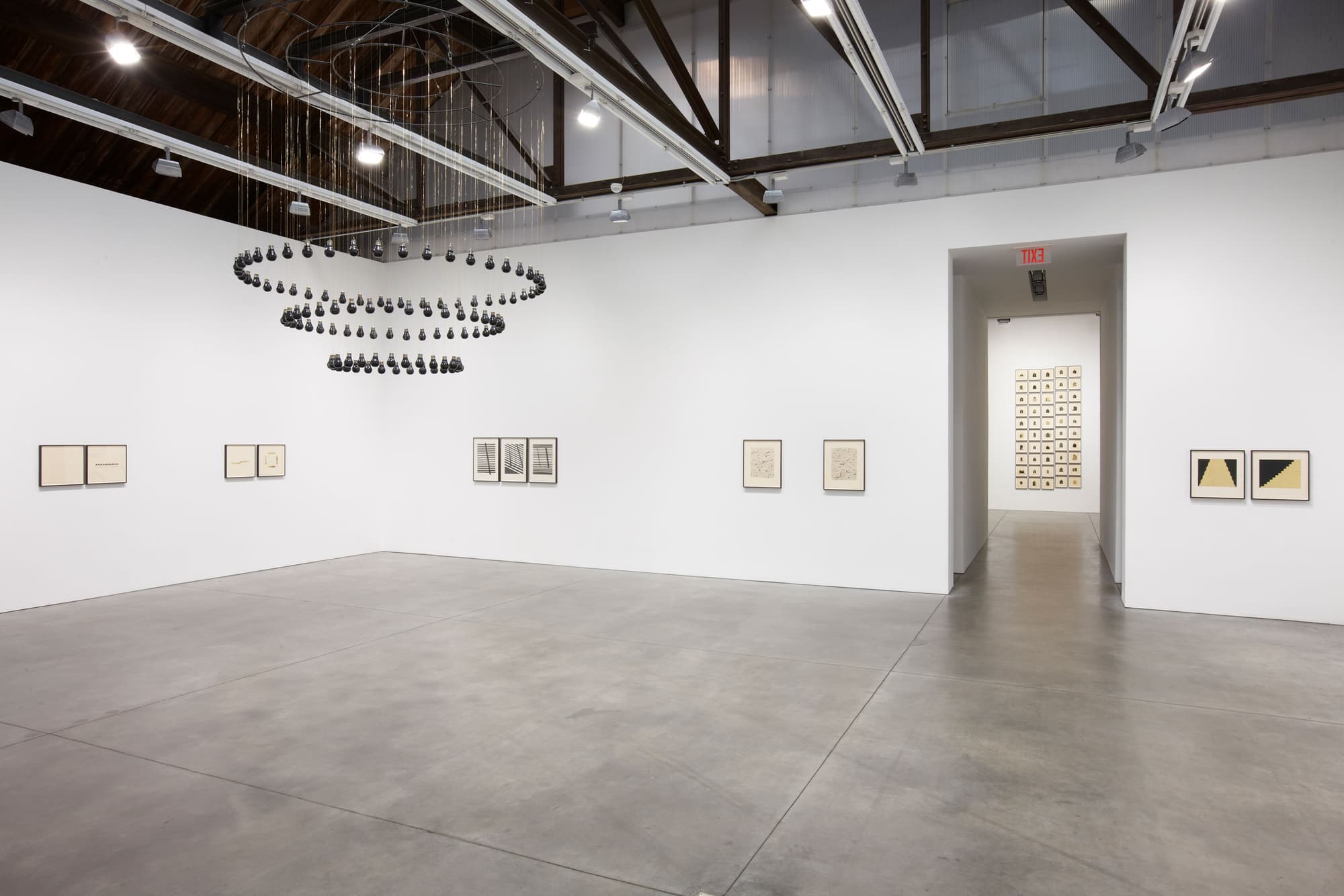 Installation view of Descending Darkness, with the suspended black-marble light bulbs of the title sculpture above works on paper